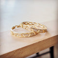 A pair of gold-tone hoop earrings featuring a geometric pattern and embedded crystals resting on a light wooden surface.