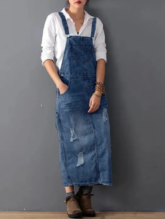 Woman wearing a distressed blue denim overall midi dress with adjustable shoulder straps, a front bib pocket, and ripped details, paired with a white long-sleeve shirt and brown boots.
