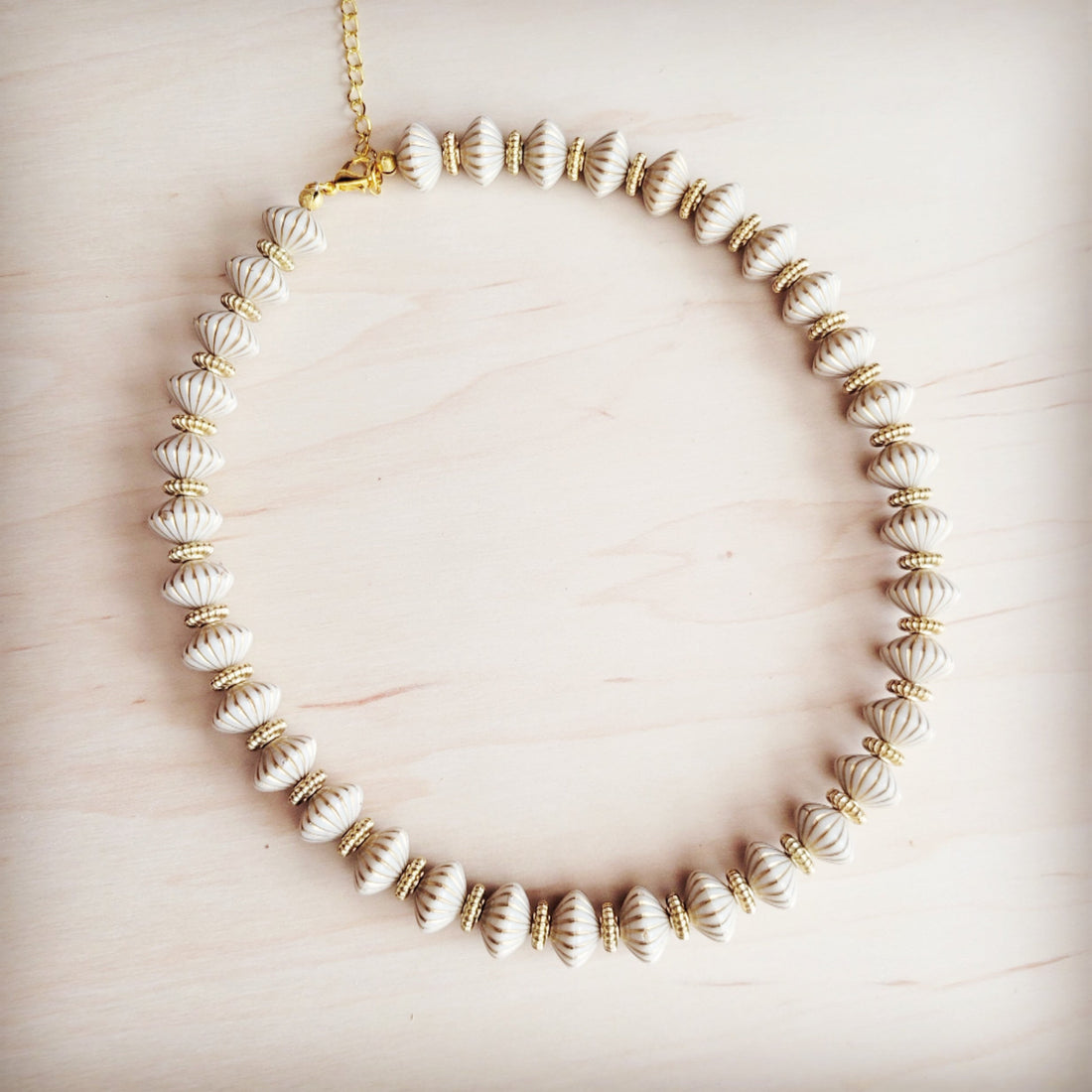 A close-up view of a creamy white, shell-textured bead collar necklace with gold-tone spacers on a light wood background.