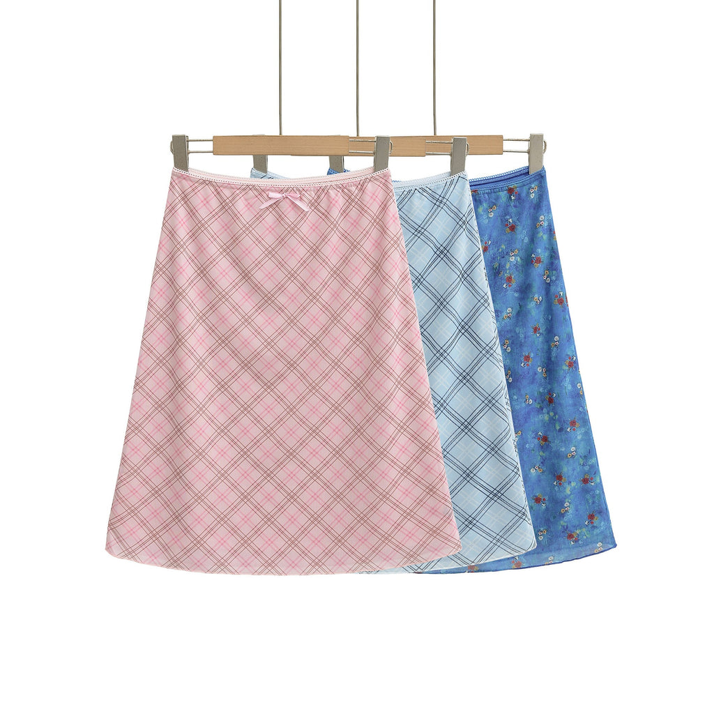 Three women's hip-hugging half skirts with elastic waists and small bows. One pink plaid skirt, one light blue plaid skirt, and one blue floral skirt, all hanging on hangers against a white background.