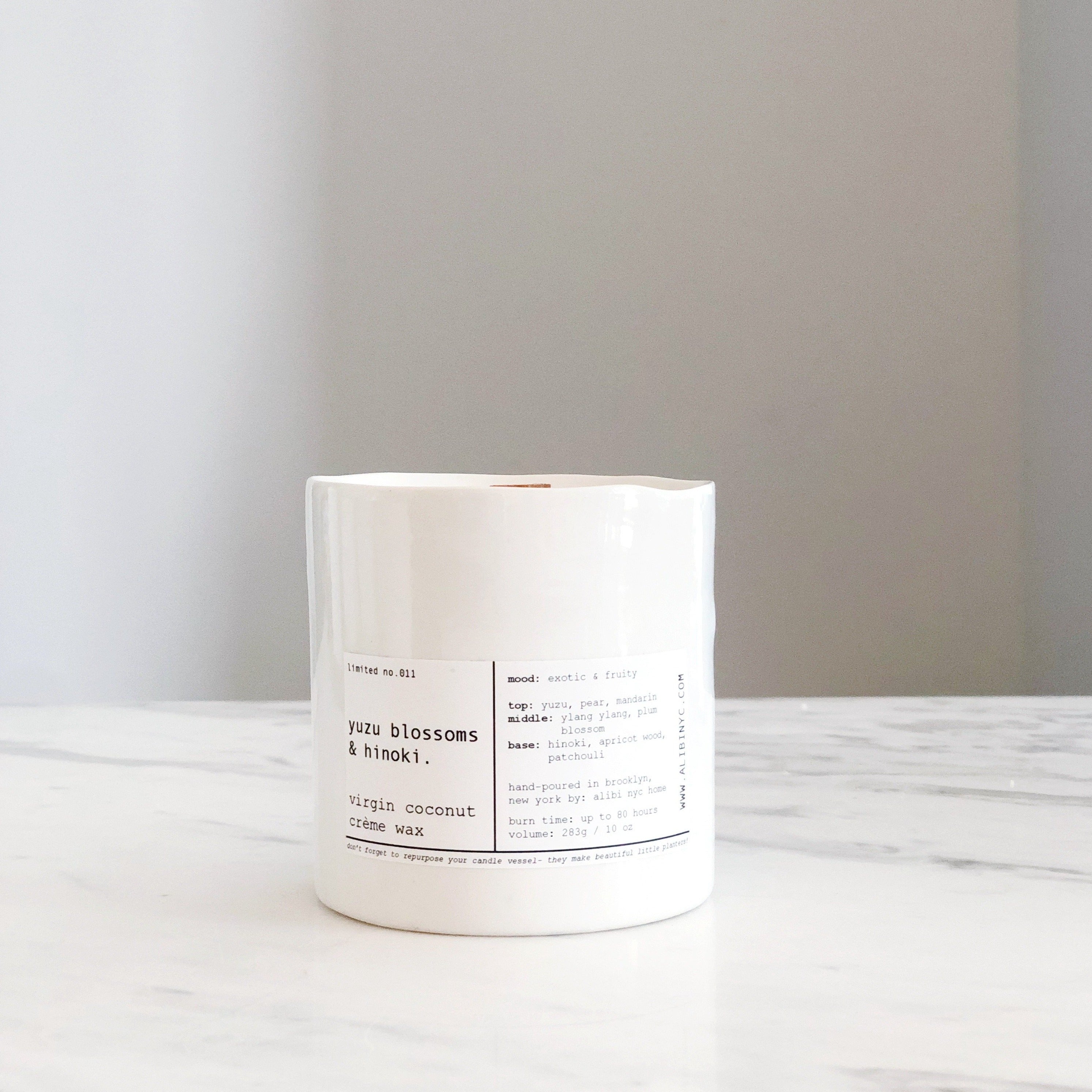 A minimalist white candle jar with a label reading Yuzu Blossom & Hinoki sitting on a white marble surface
