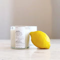 A clear glass jar candle labeled Yuzu sitting next to a fresh yellow lemon on a marble surface.