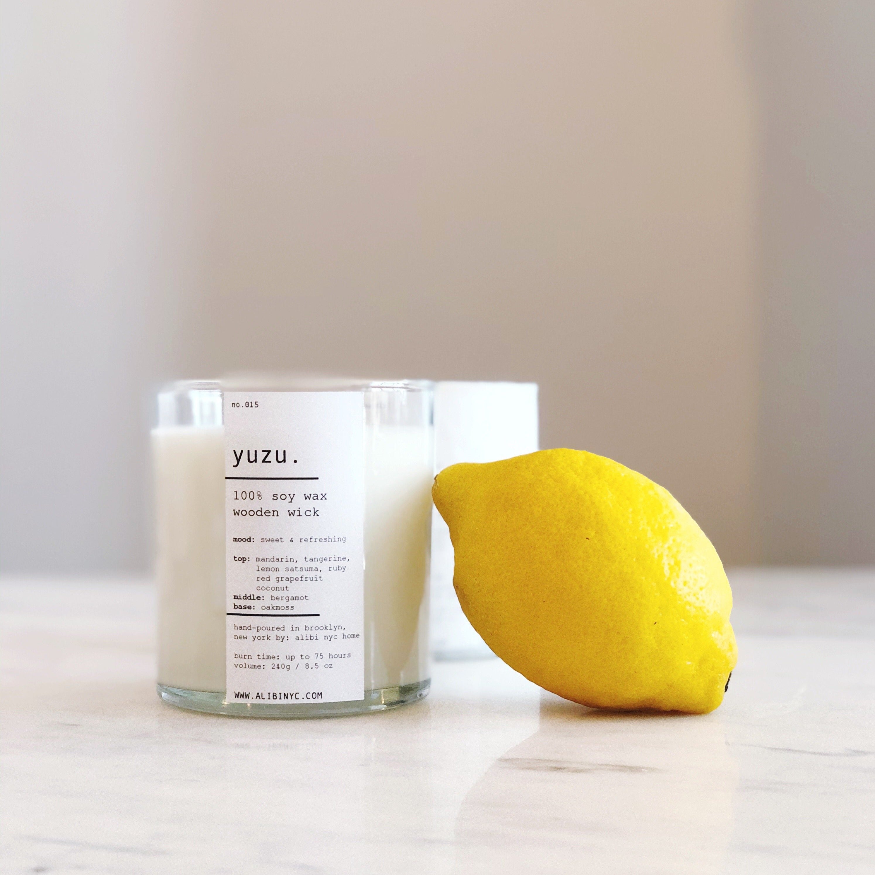 A clear glass jar candle labeled Yuzu sitting next to a fresh yellow lemon on a marble surface.