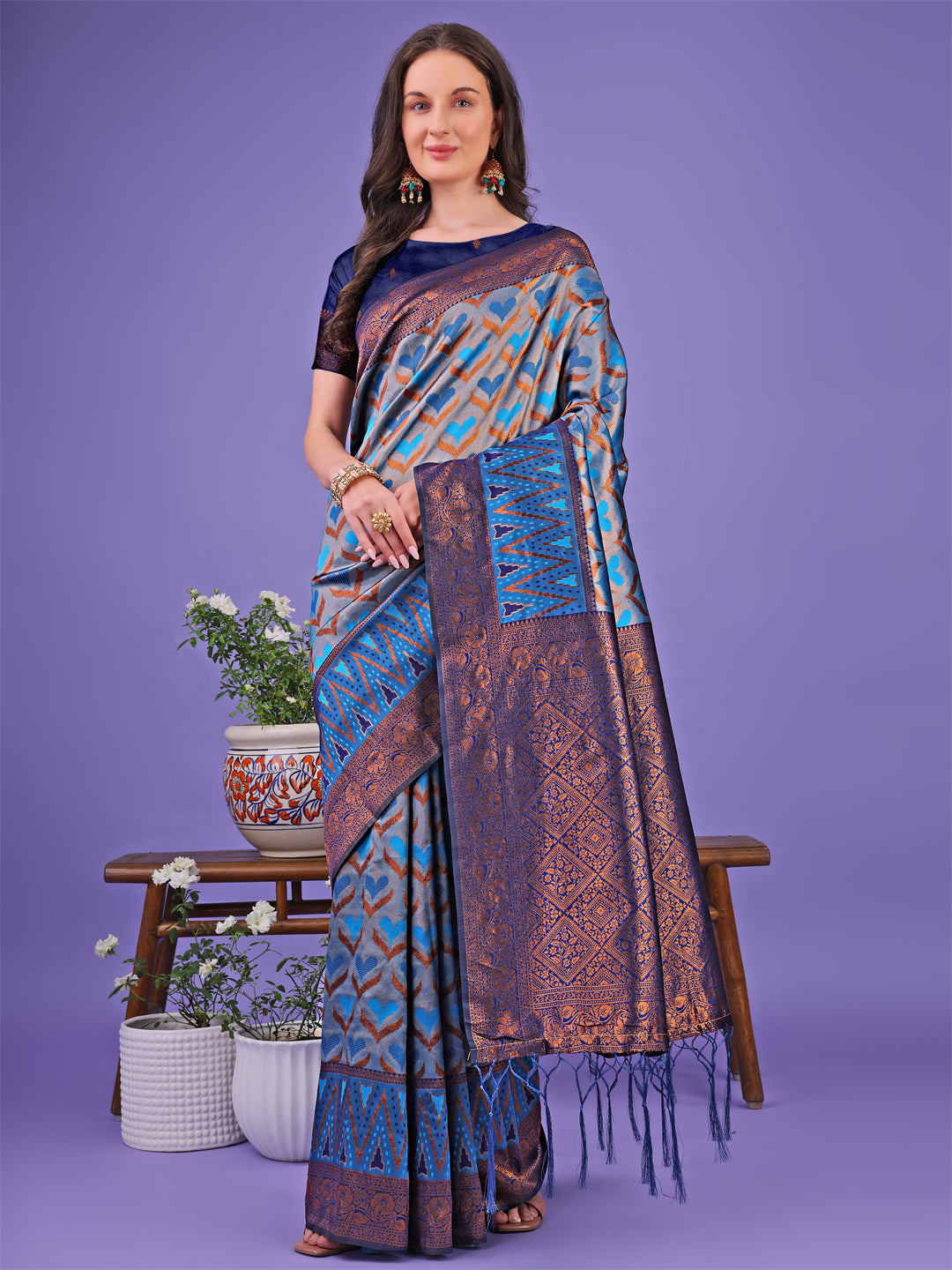Woman wearing a Radiant Lichi Silk Saree with geometric patterns in blue, copper, and grey shades.