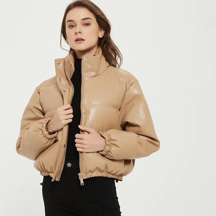 Woman wearing a tan faux leather cropped puffer jacket with a stand collar over a black top and pants, against a white background.