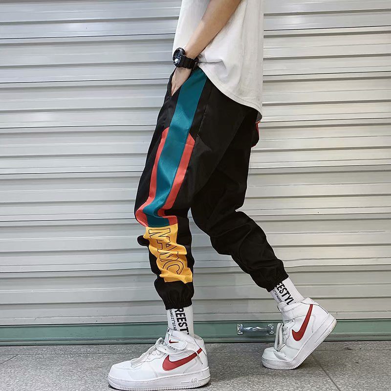Model wearing black Harajuku streetwear joggers with red, teal, and yellow side stripes and text print, styled with a white t-shirt and sneakers.