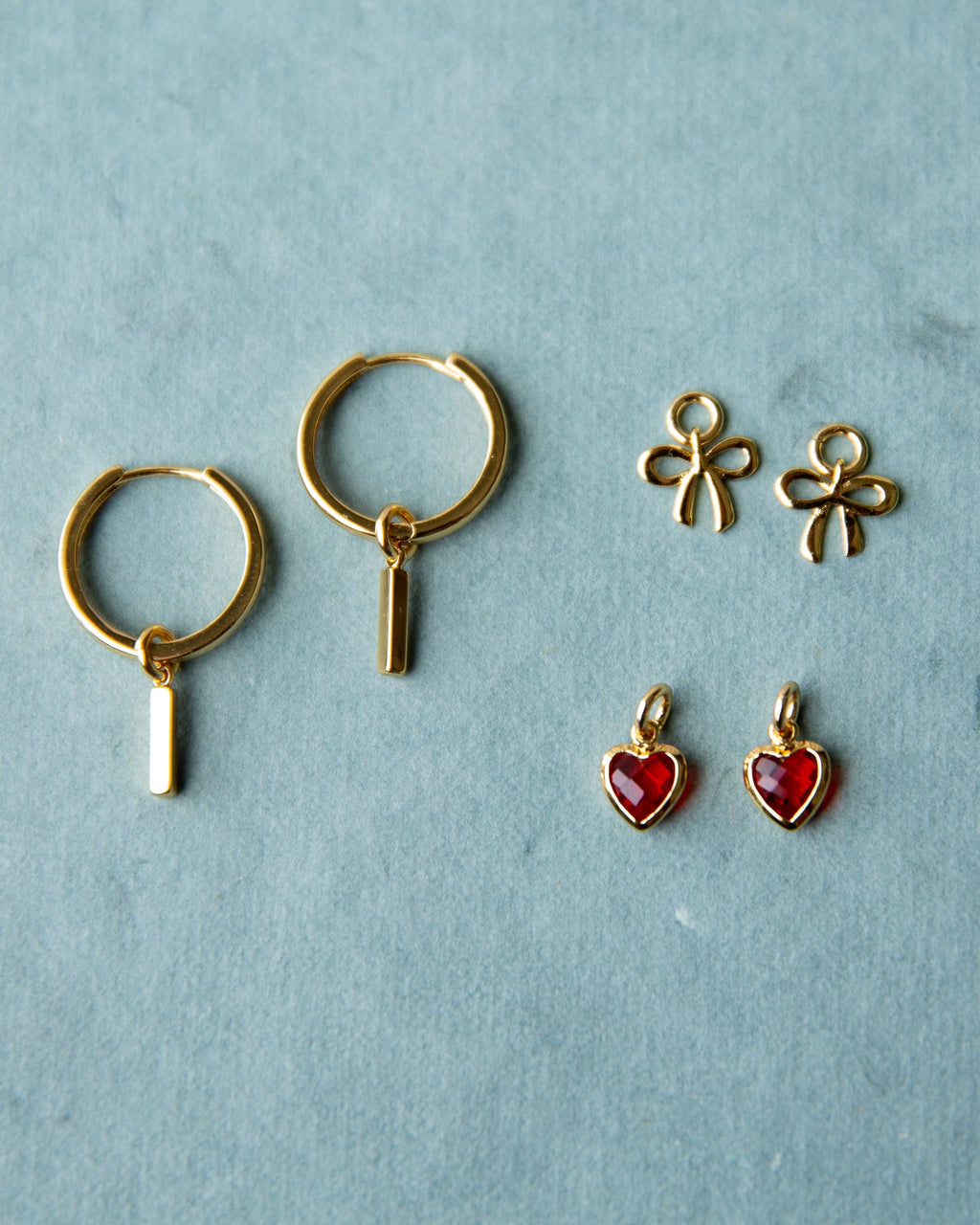 Gold huggie hoop earrings with interchangeable red heart, bow, and bar charms on a white background.
