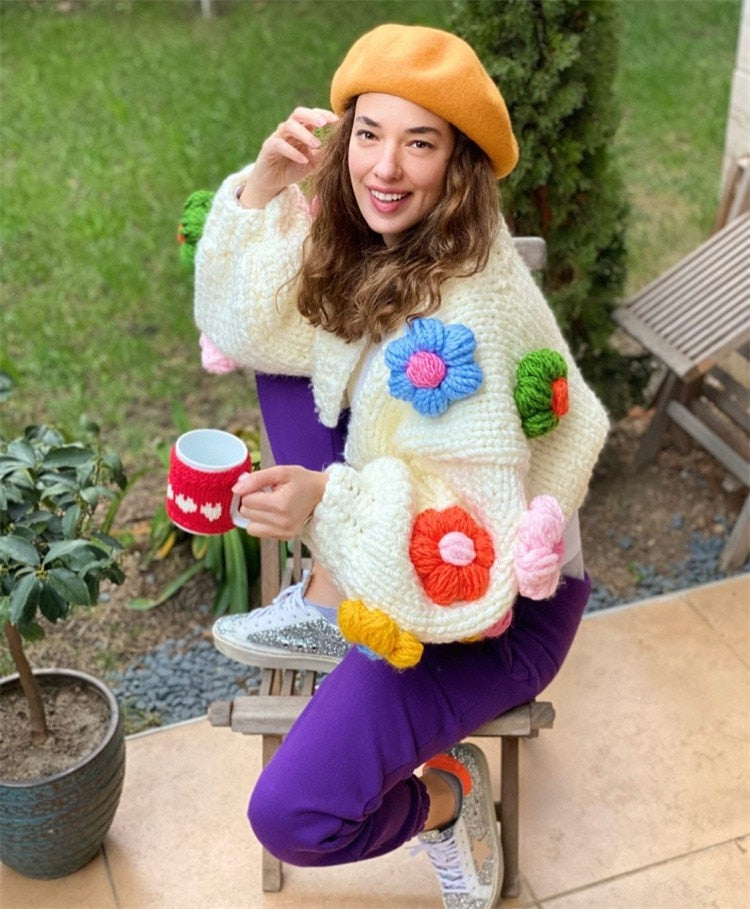 Woman wearing a cream knit cardigan with colorful 3D flower appliques and a yellow beret.