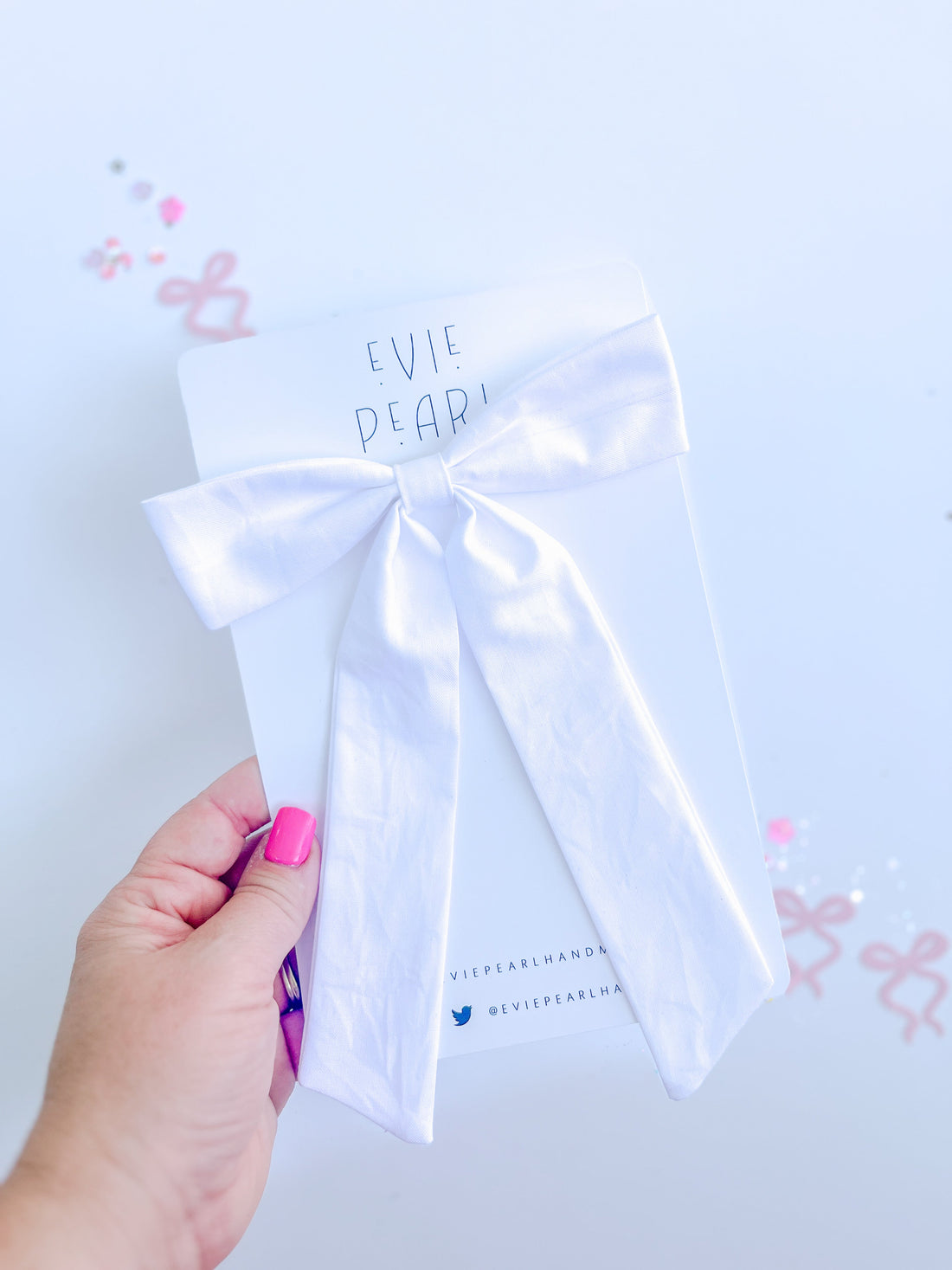 A white long-tailed fabric hair bow attached to a branded card held by a hand.