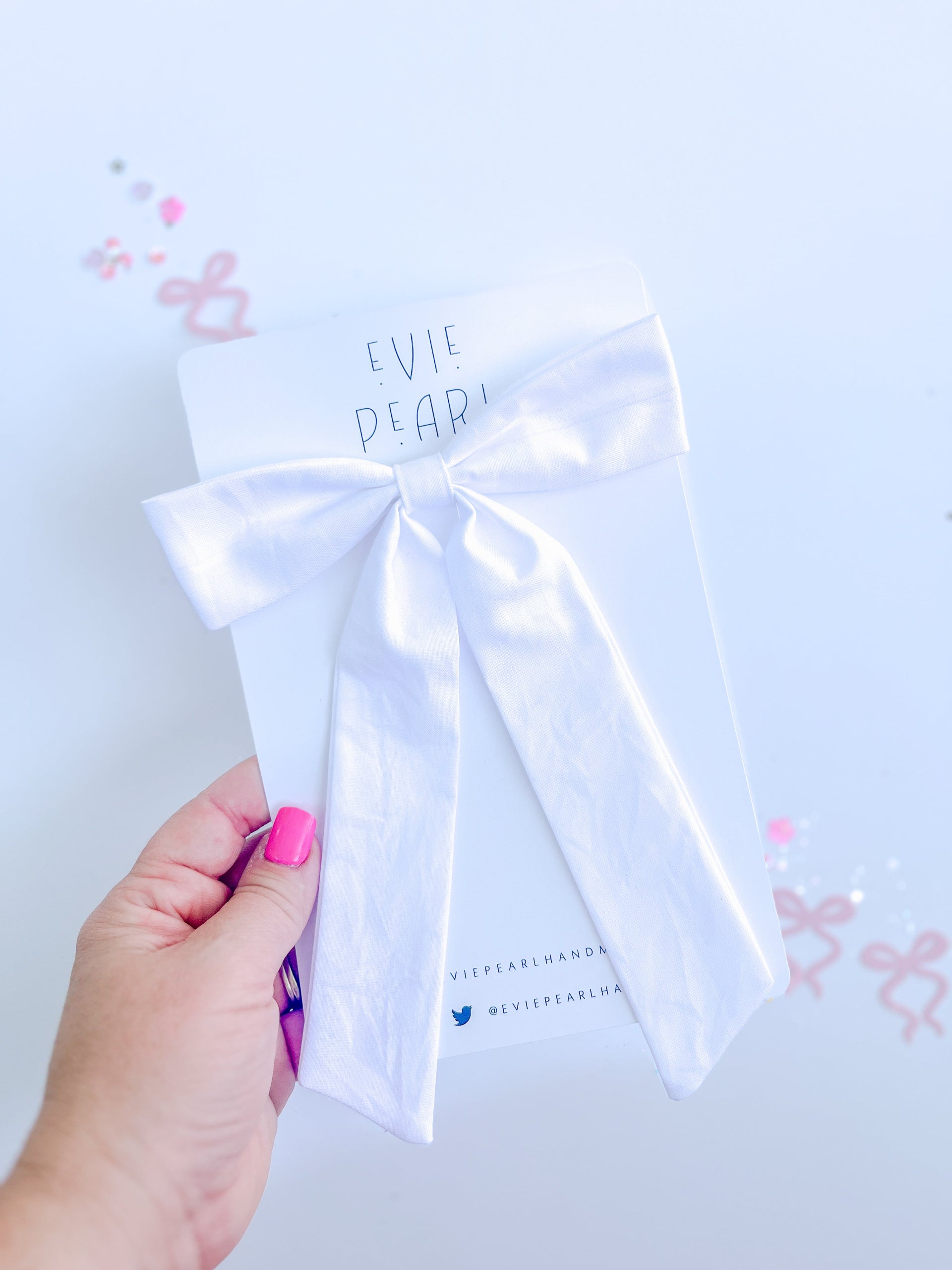 A white long-tailed fabric hair bow attached to a branded card held by a hand.