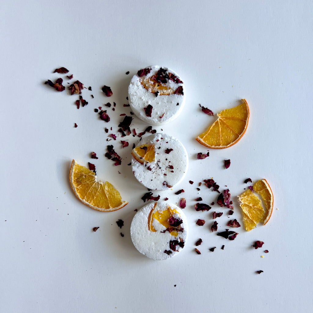 Three round white shower steamers decorated with dried orange slices and rose petals arranged on a white surface.