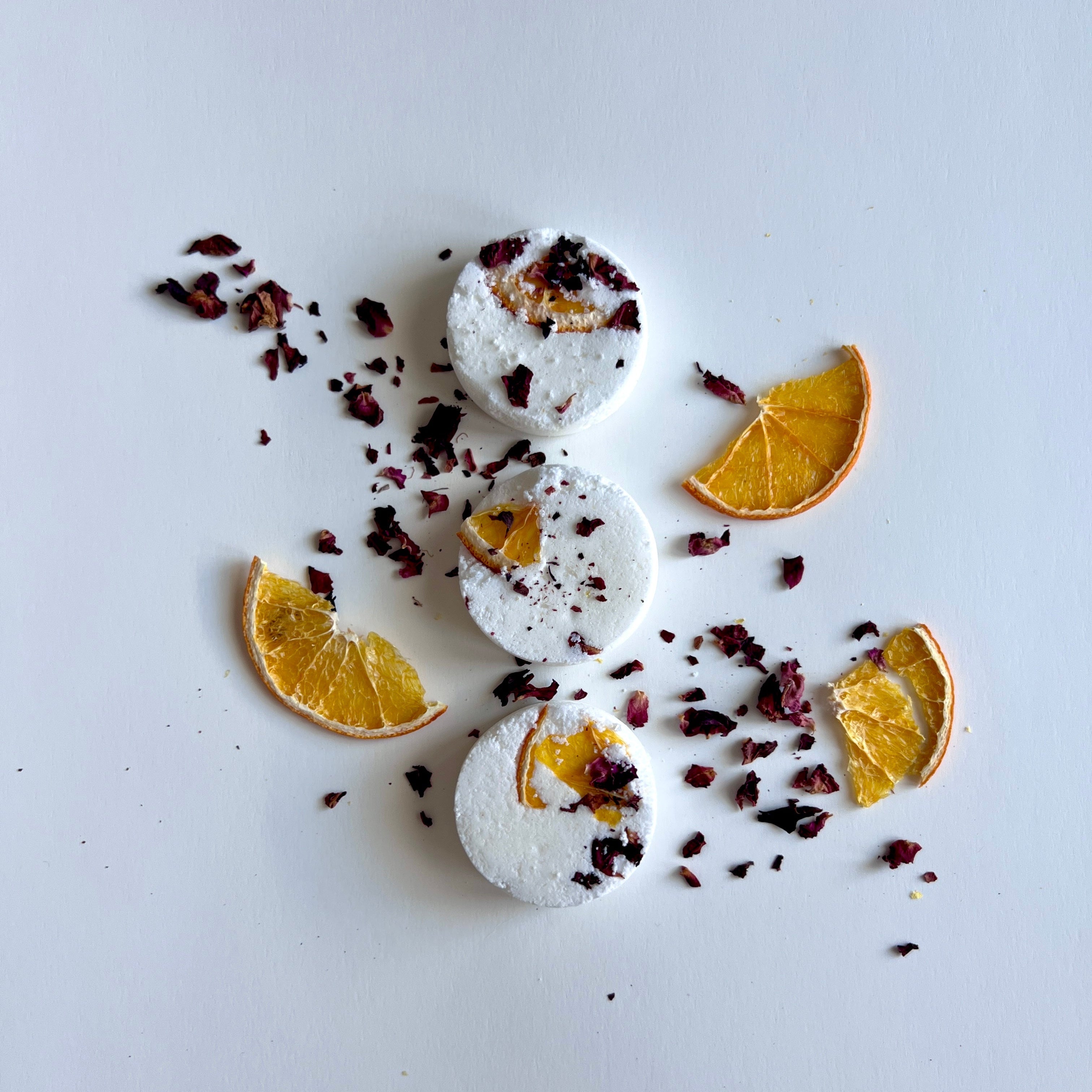 Three round white shower steamers decorated with dried orange slices and rose petals arranged on a white surface.