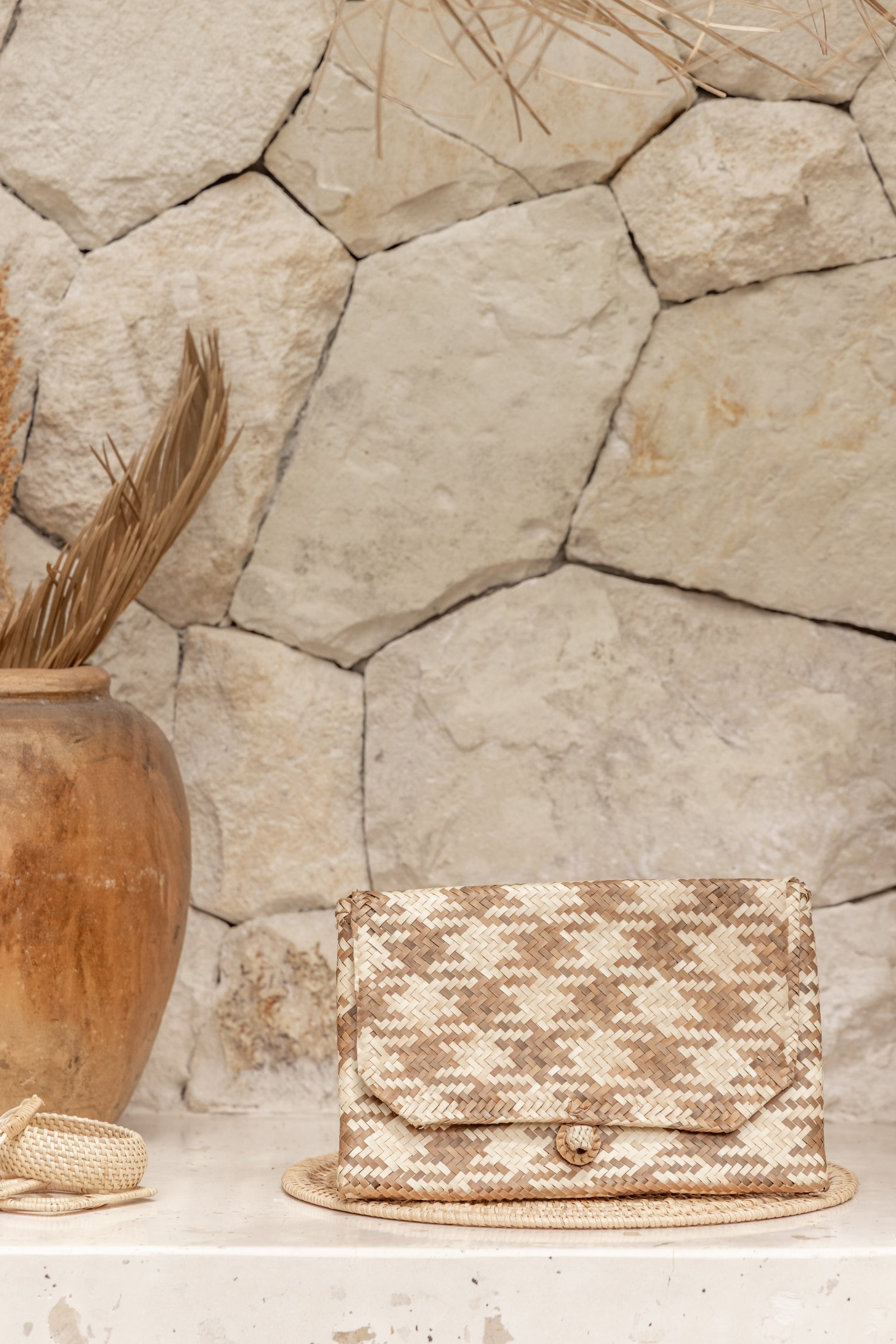 A woven rattan clutch with a plaid pattern sitting on a stone surface next to a terracotta vase.