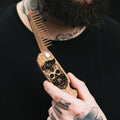 Close-up of a handmade natural walnut wooden beard comb with a laser-engraved skull design held by a man.