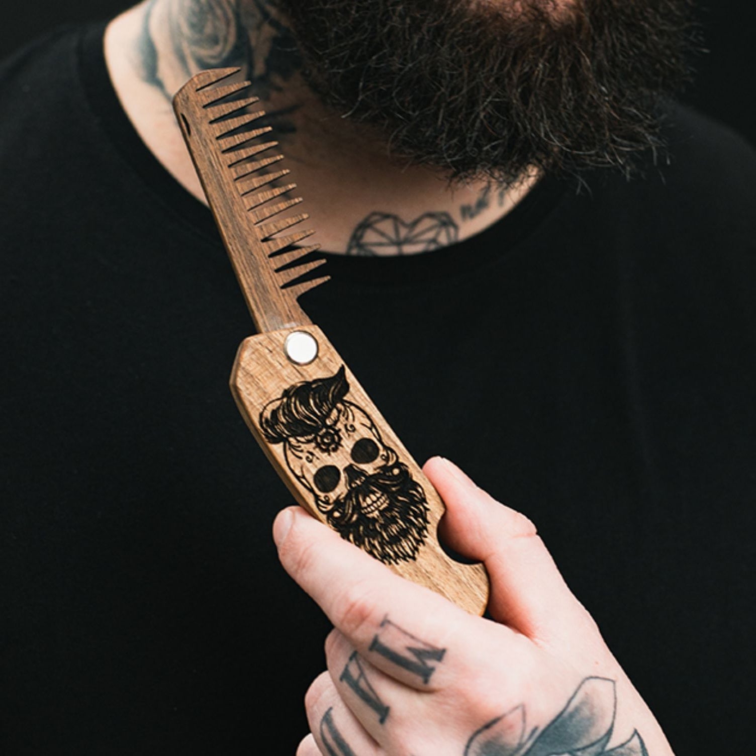 Close-up of a handmade natural walnut wooden beard comb with a laser-engraved skull design held by a man.