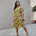 A woman wearing a vibrant yellow floral midi dress with a V-neck, short sleeves, and a smocked waist in an outdoor setting.
