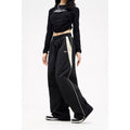 Woman wearing black Harajuku style loose wide-leg track pants with a beige side stripe and drawstring waist. Another image shows brown wide-leg pants with a black stripe.