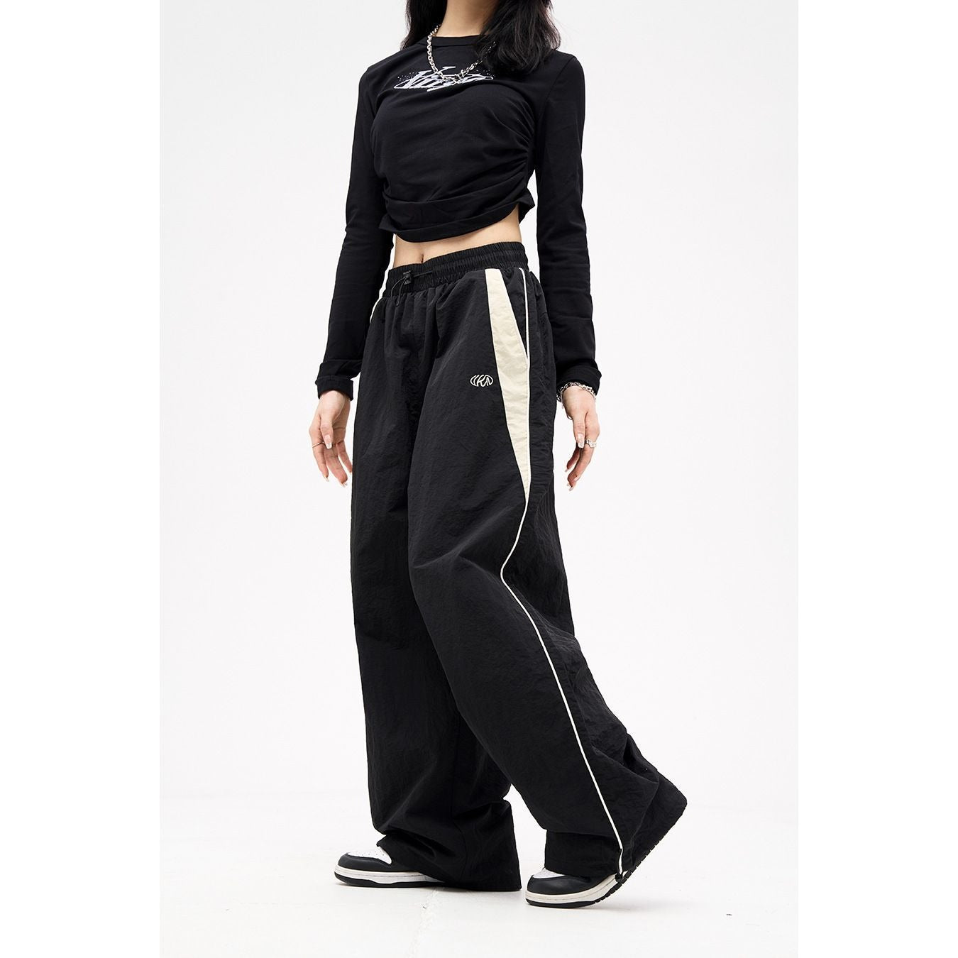 Woman wearing black Harajuku style loose wide-leg track pants with a beige side stripe and drawstring waist. Another image shows brown wide-leg pants with a black stripe.