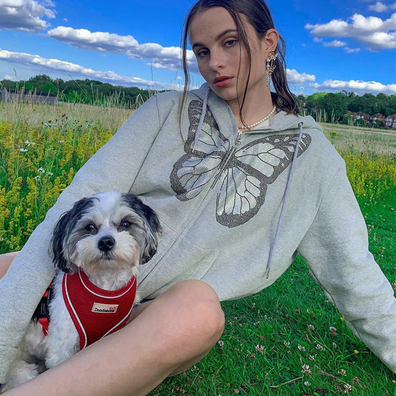 A woman wearing a grey zip-up hoodie with a large, sparkly butterfly graphic, sitting in a grassy field with a small dog.