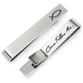 Silver tone tie bar featuring an ichthys fish symbol on the front and Come Follow Me engraved on the back, shown on a black tie.