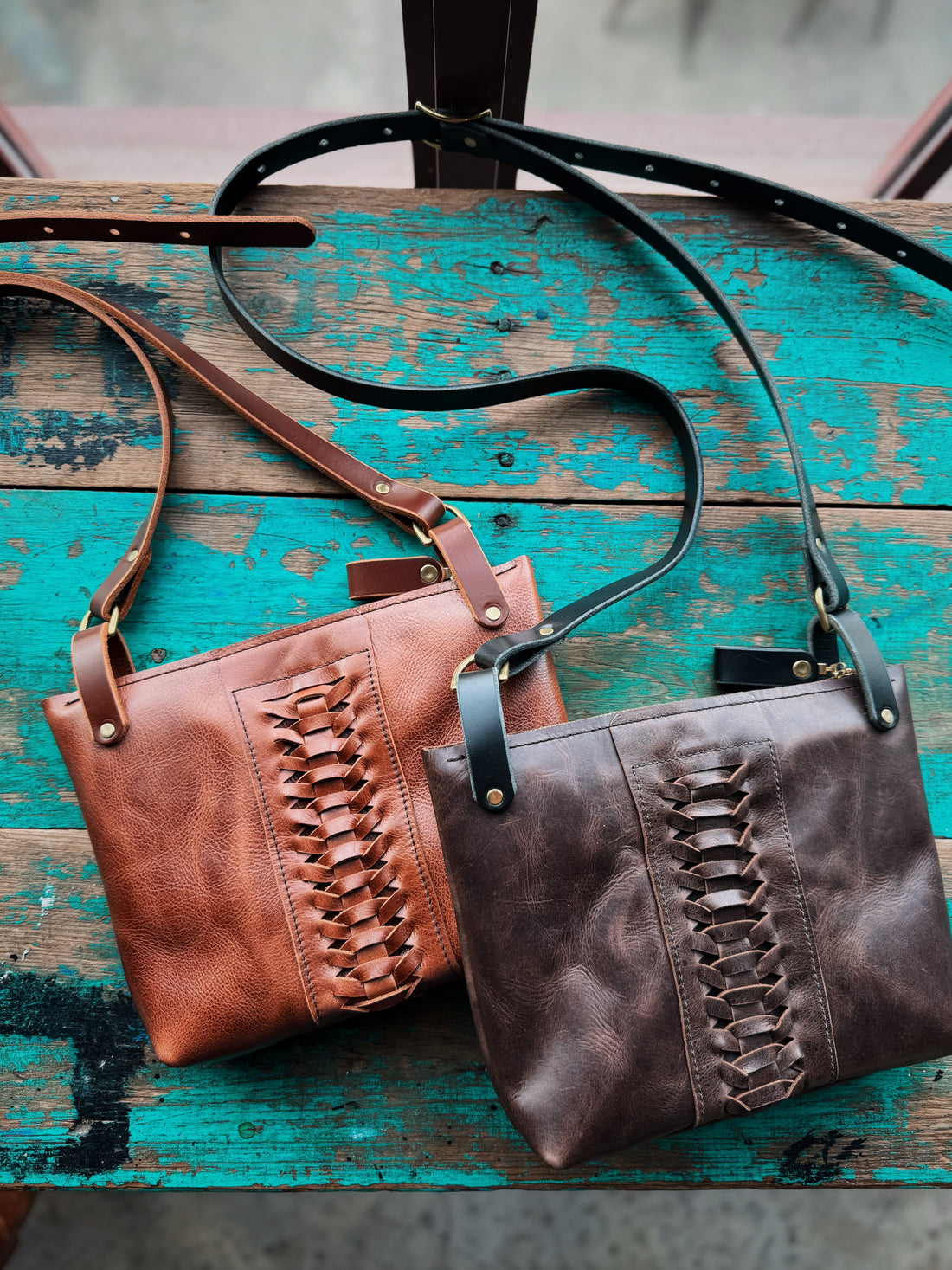Two woven leather crossbody bags, one in tan and one in dark brown, resting on a rustic teal wooden surface.