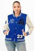 A woman wearing a royal blue varsity bomber jacket with cream sleeves and graphic patches on a white background.