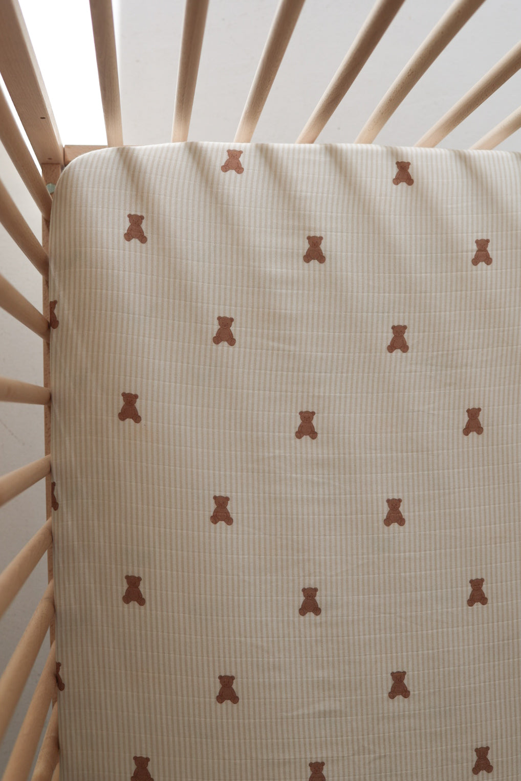 Close up and full view of a neutral striped muslin crib sheet with small brown teddy bear print inside a wooden crib.