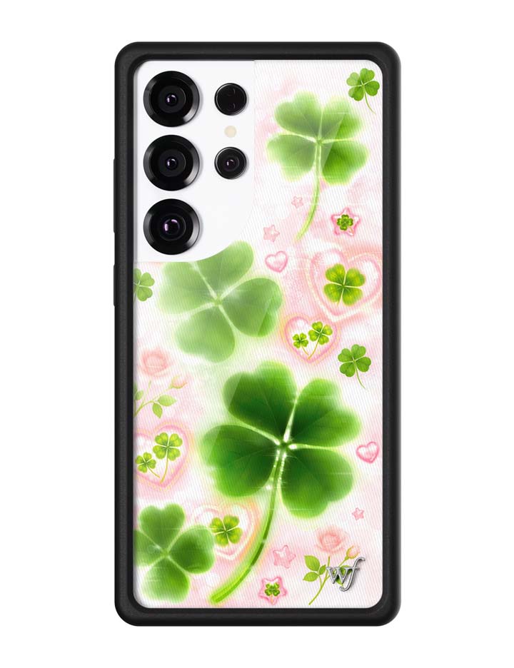 Samsung Galaxy phone case featuring a light background with green four-leaf clovers, pink hearts, small stars, and rose illustrations, encased in a black rubber bumper.