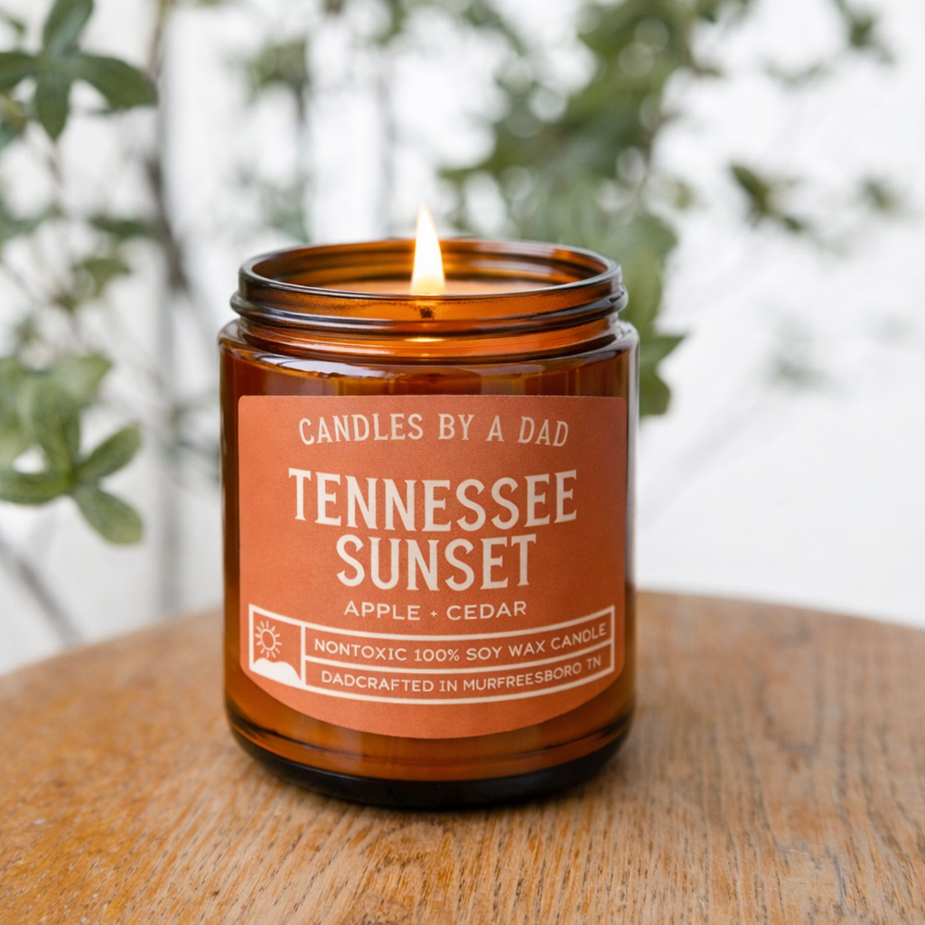 Tennessee Sunset soy wax candle in an amber glass jar on a wooden surface with greenery in the background