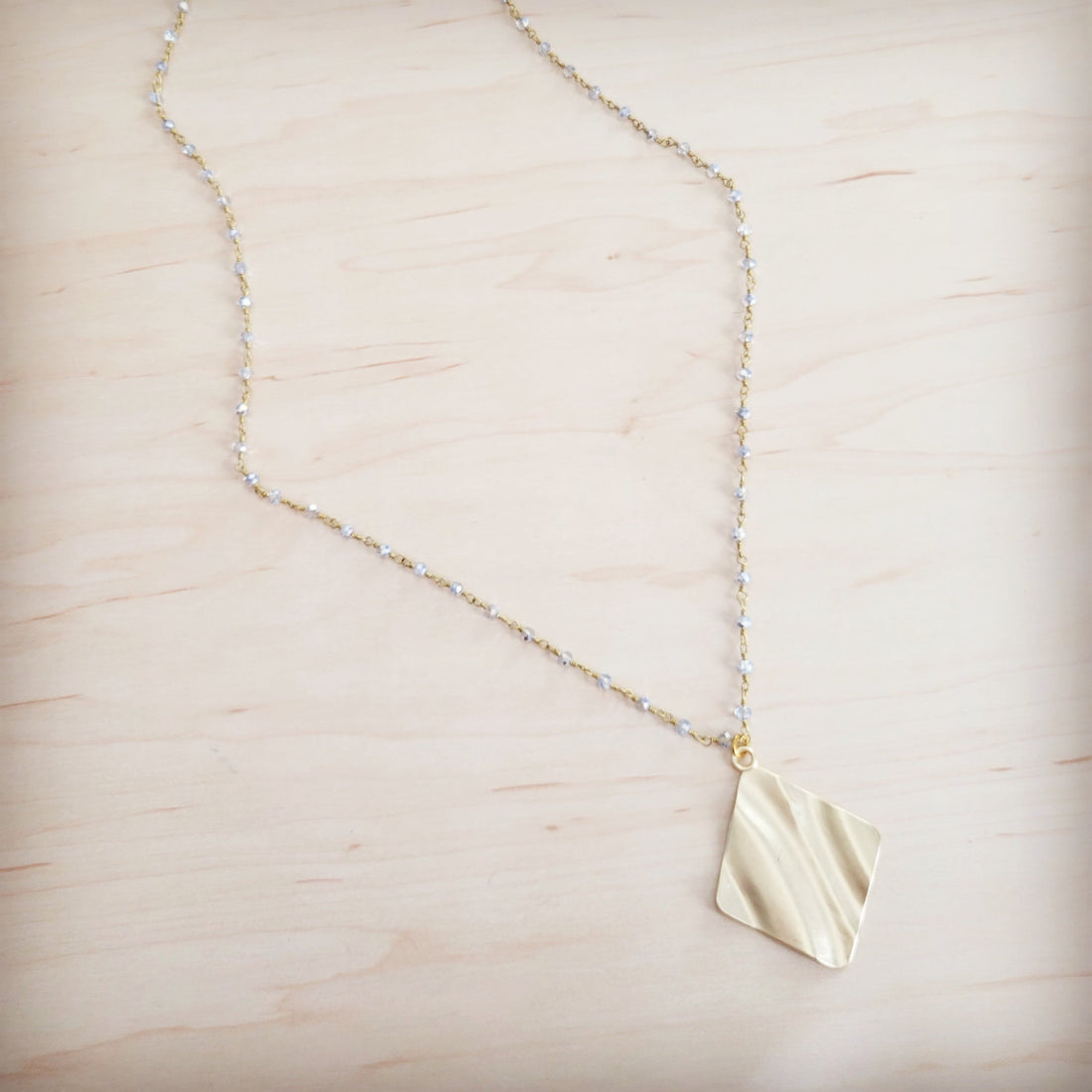 Gold-tone diamond-shaped wavy pendant necklace with a beaded chain on a light wooden background