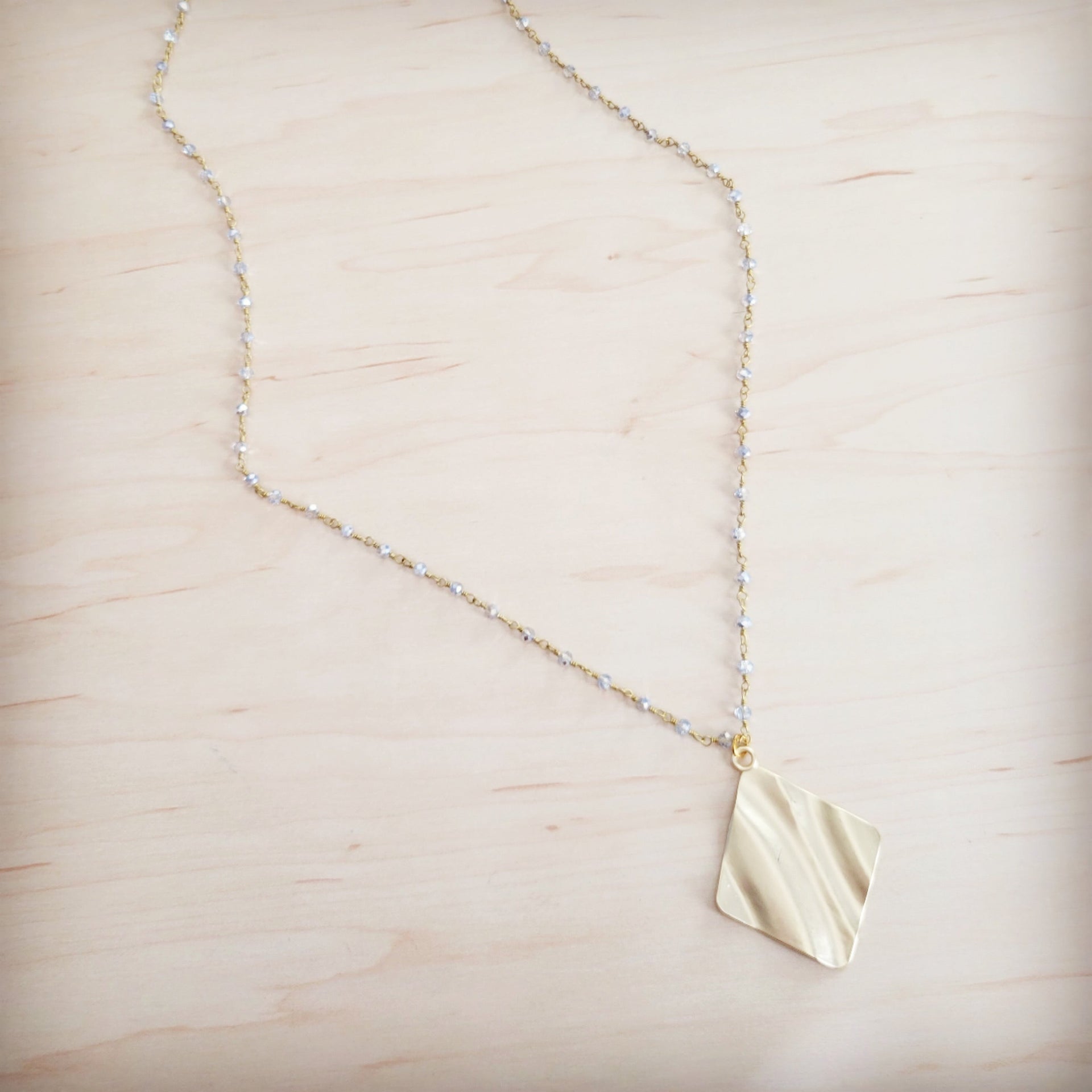 Gold-tone diamond-shaped wavy pendant necklace with a beaded chain on a light wooden background