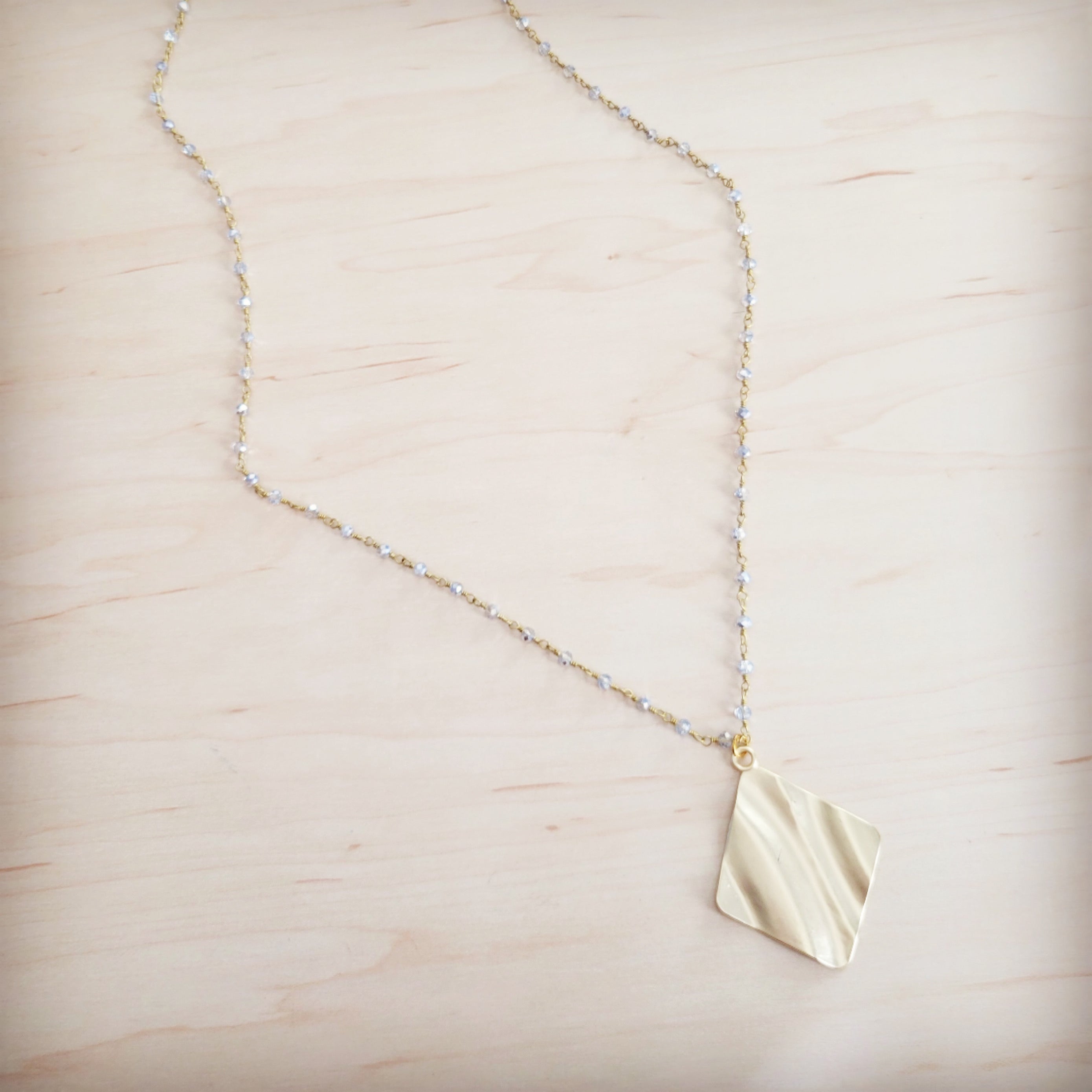 Gold-tone diamond-shaped wavy pendant necklace with a beaded chain on a light wooden background