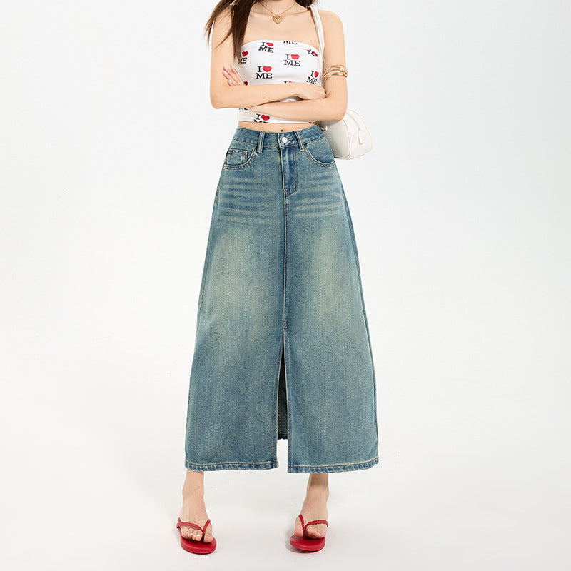 A woman wearing a high-waisted, A-line blue denim maxi skirt with a front slit, paired with a white crop top and red sandals.