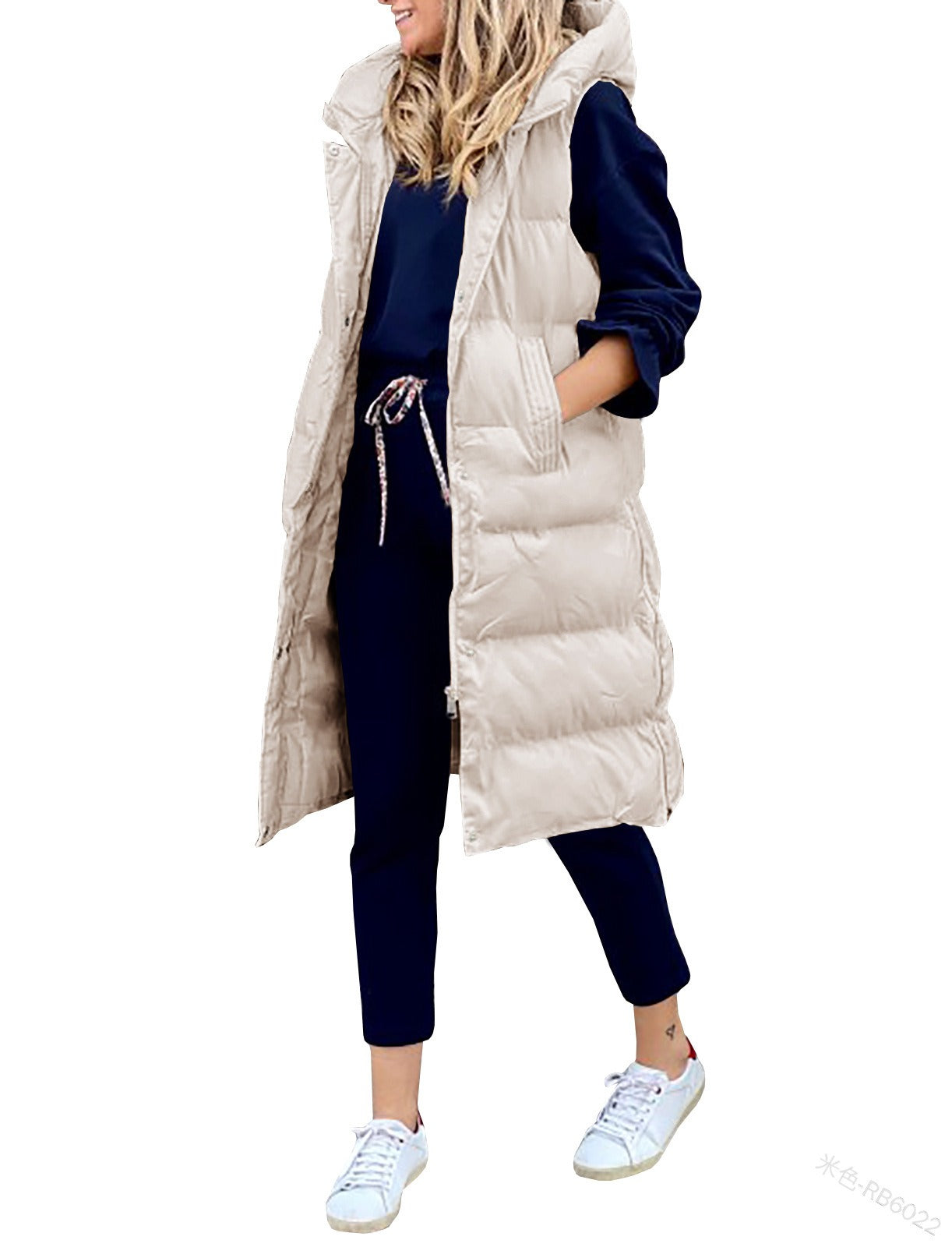 A woman wearing a long, hooded beige puffer vest layered over a dark outfit, standing in a studio setting.