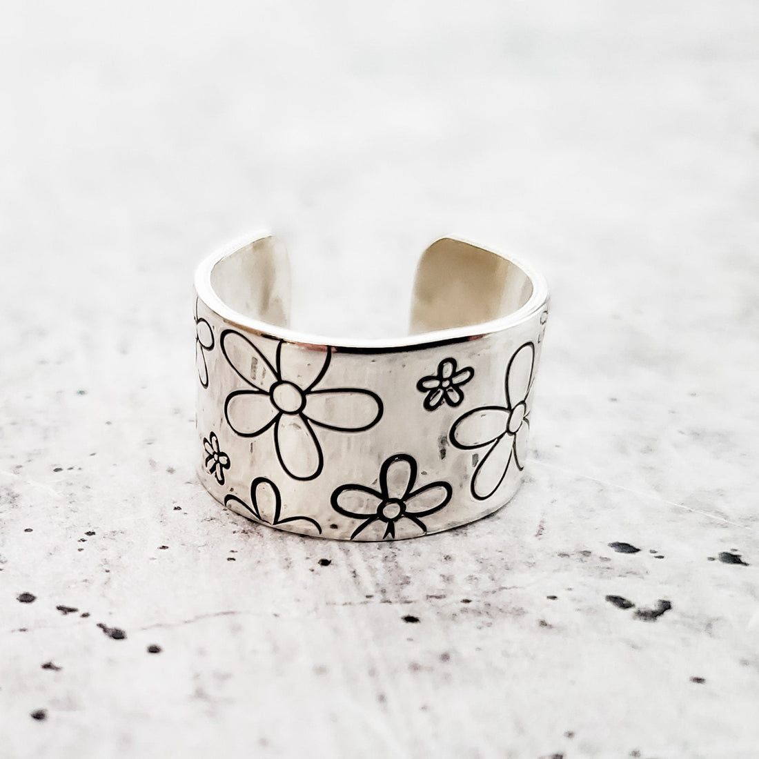 A silver-tone wide band ring featuring a stamped daisy flower pattern, shown on a textured white background.