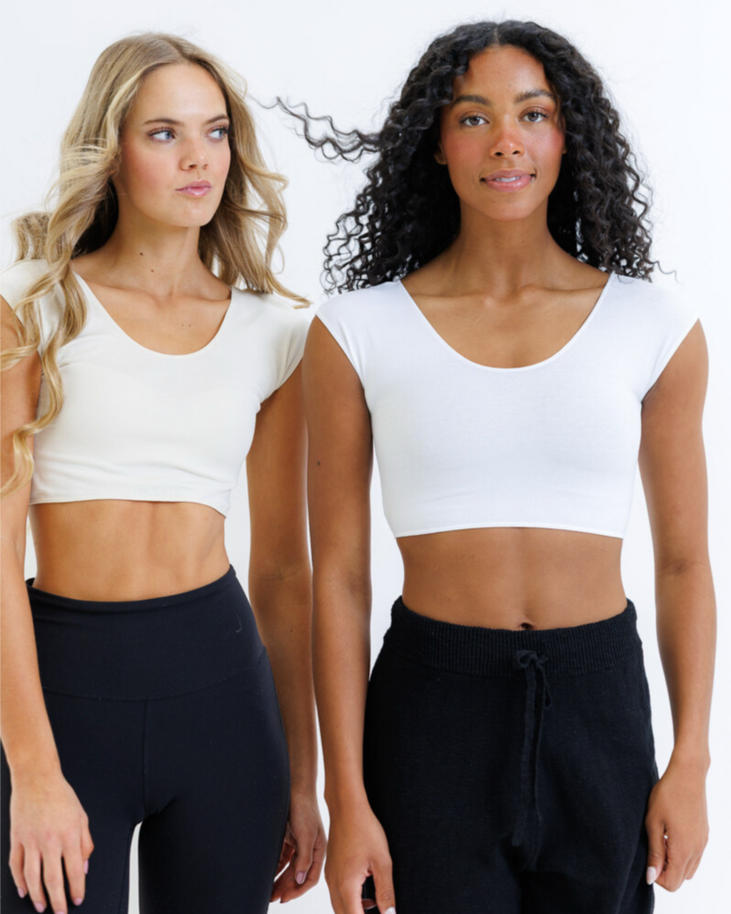 Two models wearing white Demi crop tops with scoop necks and cap sleeves paired with dark bottoms.