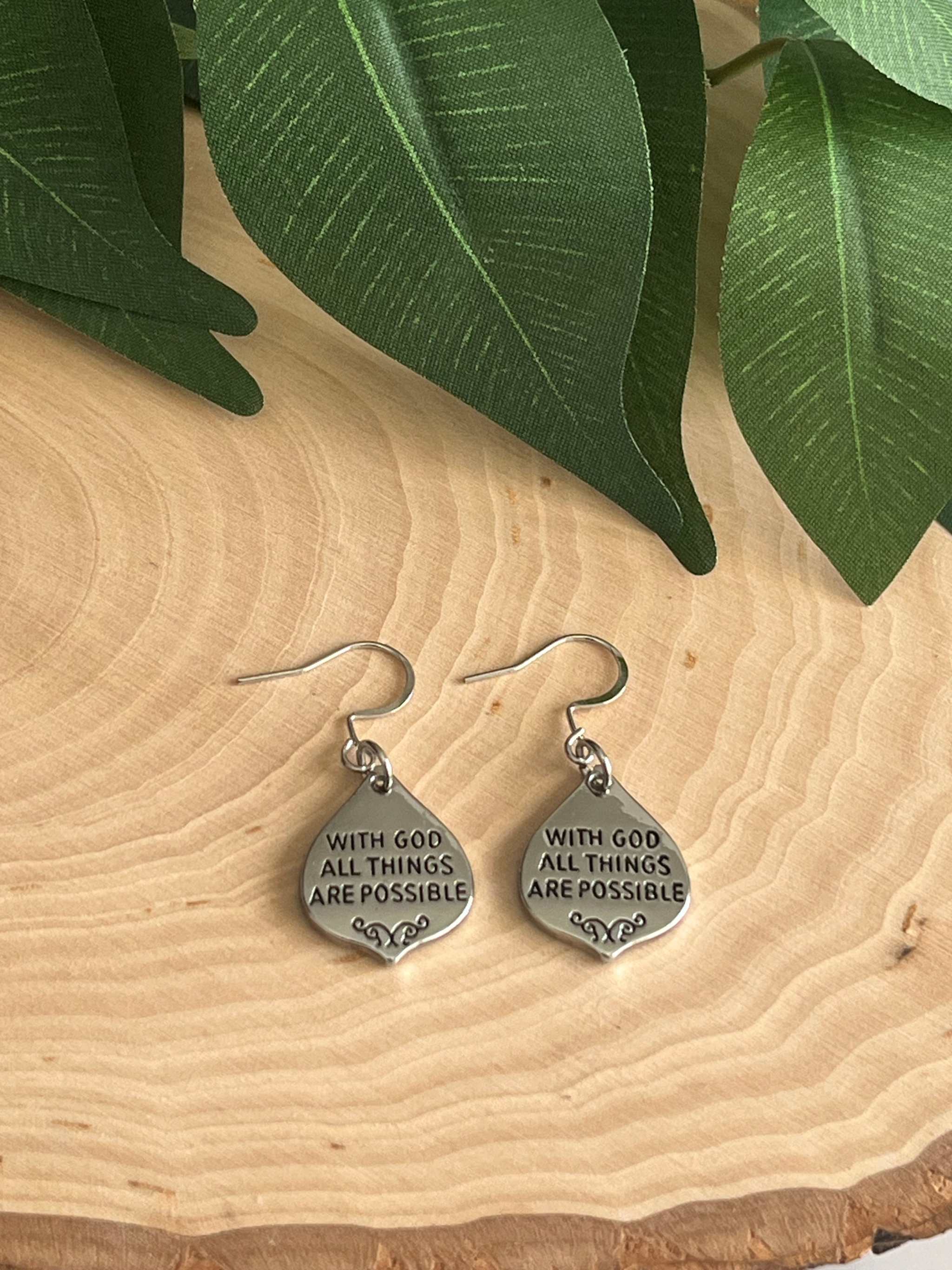 Silver drop earrings with the text With God All Things Are Possible engraved on a teardrop charm resting on a wood slice