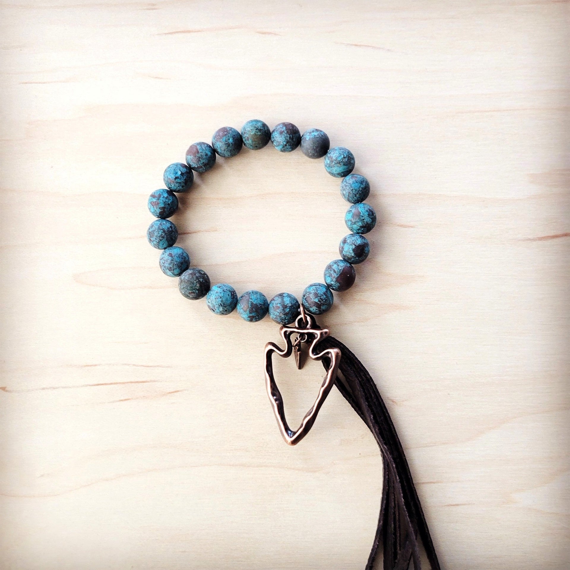 Turquoise bead stretch bracelet with copper-tone arrowhead charm and leather-style tassel on wood background