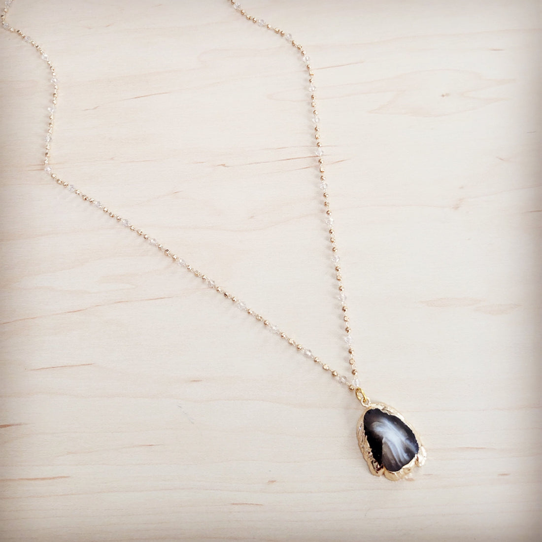 A close-up shot of a black agate pendant necklace with a gold-tone trim on a delicate beaded chain resting on a light wood background.