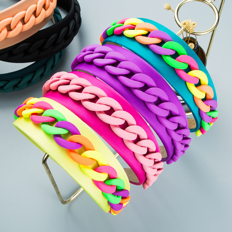 Collection of wide fabric headbands in various vibrant macaron colors (pink, purple, blue, yellow, teal) each adorned with a multi-colored plastic chain detail, displayed on a gold stand.
