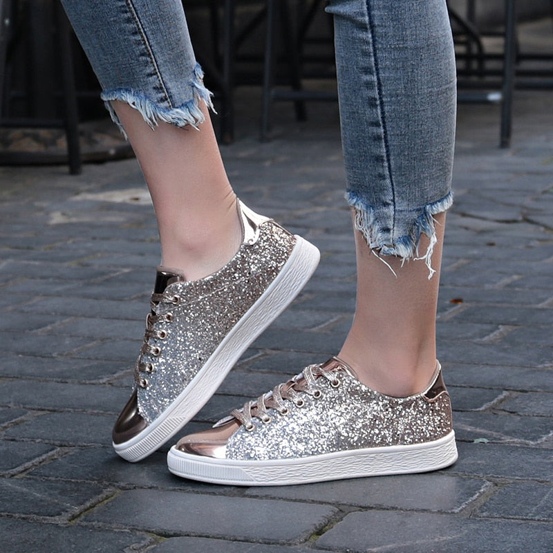 A woman wearing Bling Green glitter metallic sneakers with ripped jeans. The shoes feature a sparkling green upper with a shiny metallic toe cap and heel, and white rubber soles.