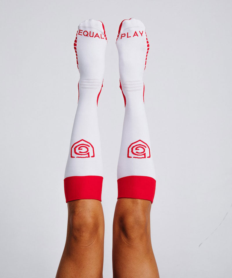 A pair of white over-the-calf soccer socks with red cuffs and red branding detail worn by a female athlete.