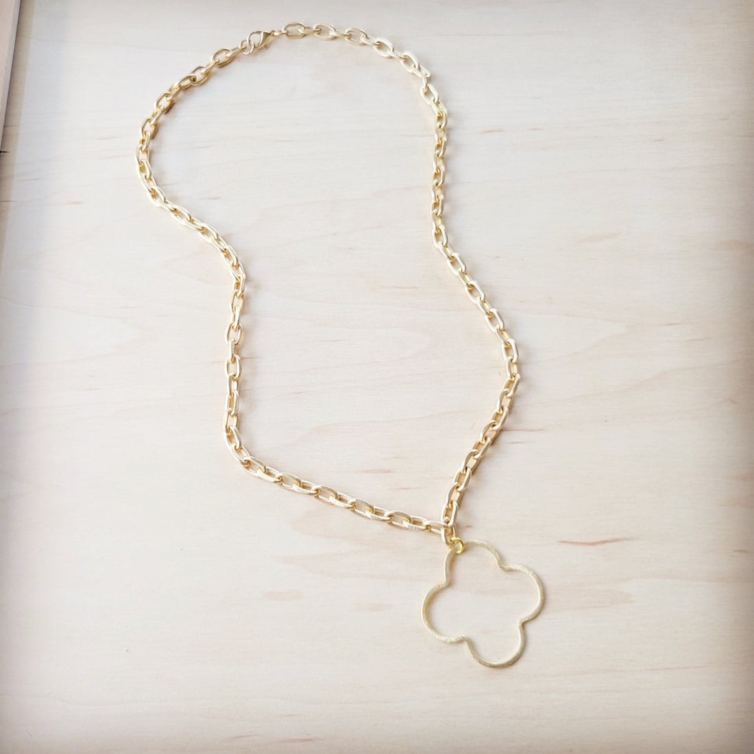 A matte gold-tone necklace with an open quatrefoil pendant hanging from an oval-link chain on a light wooden background.