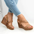 A pair of casual tan wedge sandals with a cross-strap design being worn with blue jeans.