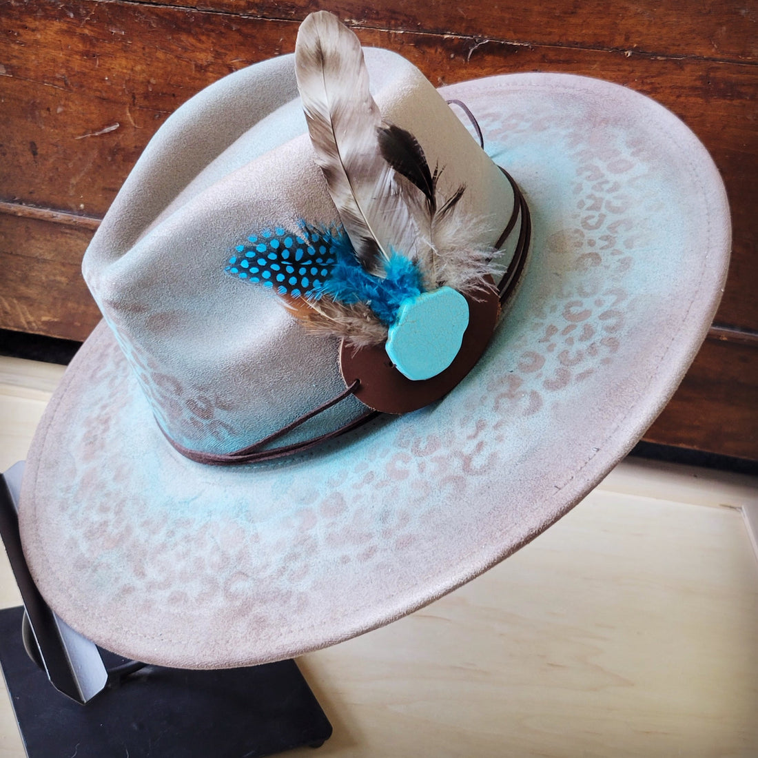 Hand-painted tan cowgirl hat with leopard print, brown leather band, turquoise stone, and blue and brown feathers.