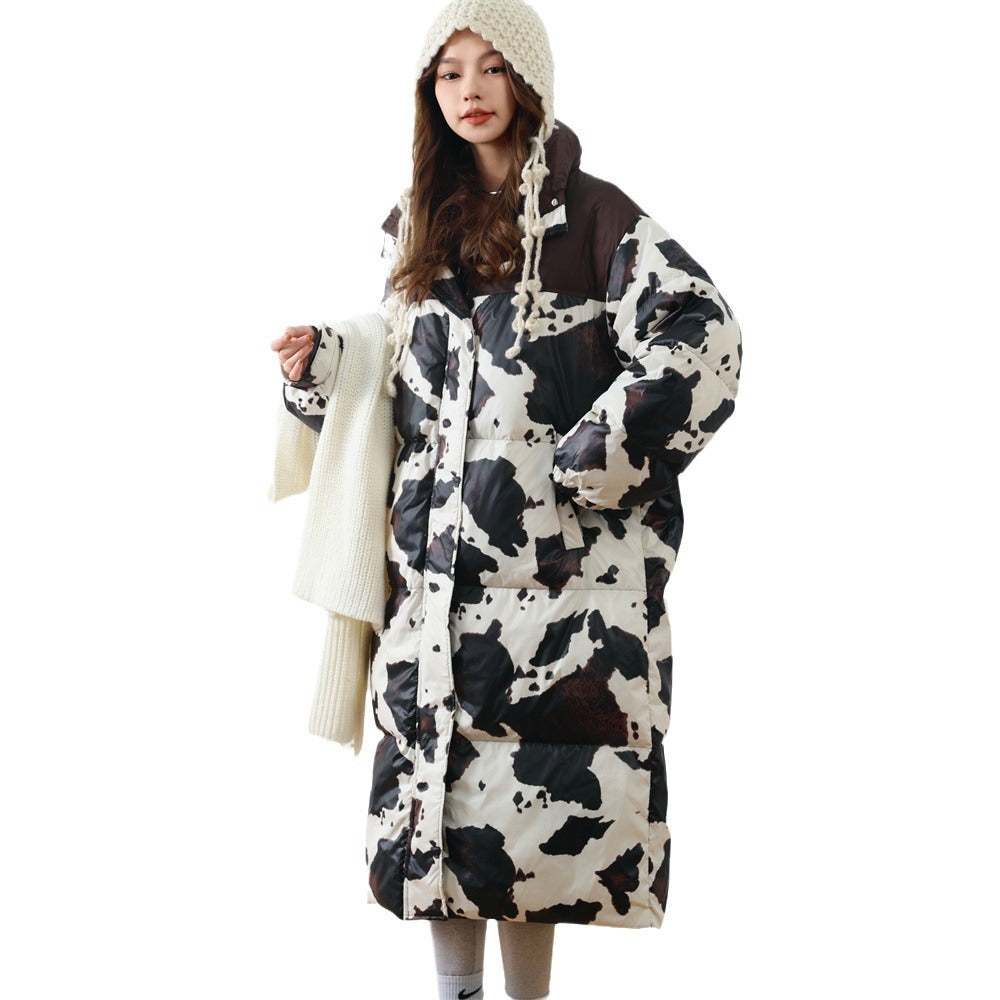 Woman wearing a long Harajuku style cow print white and black duck down puffer coat with a white knitted hat and scarf, standing against a white background.