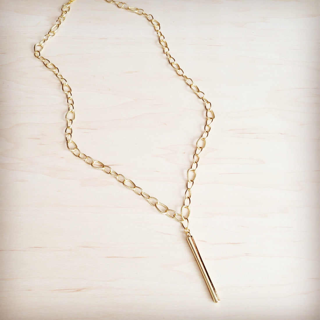 A gold-tone long chain necklace with a sleek vertical bar pendant resting on a light wooden surface.