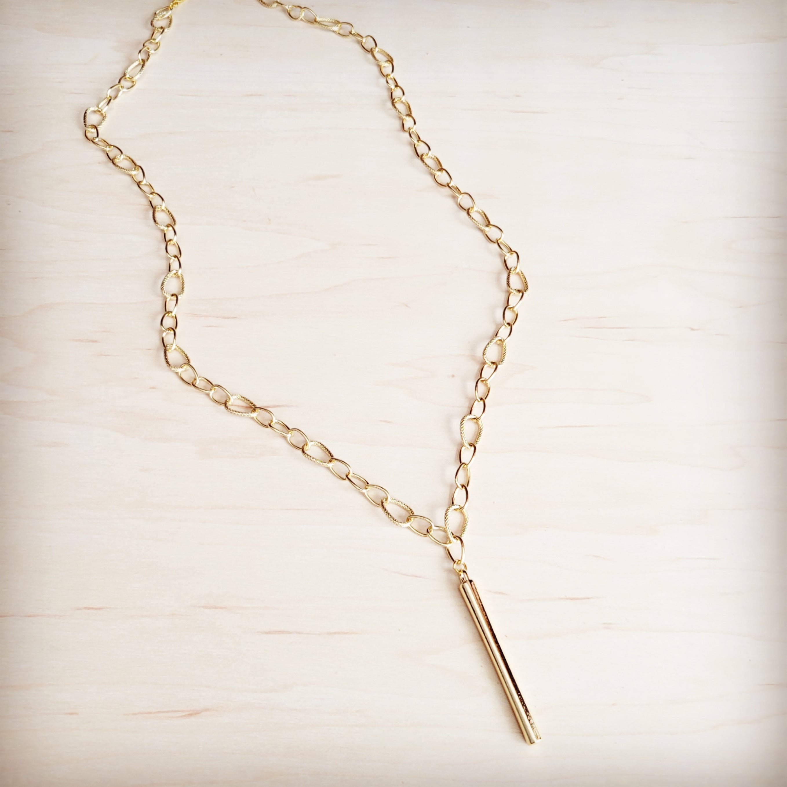 A gold-tone long chain necklace with a sleek vertical bar pendant resting on a light wooden surface.