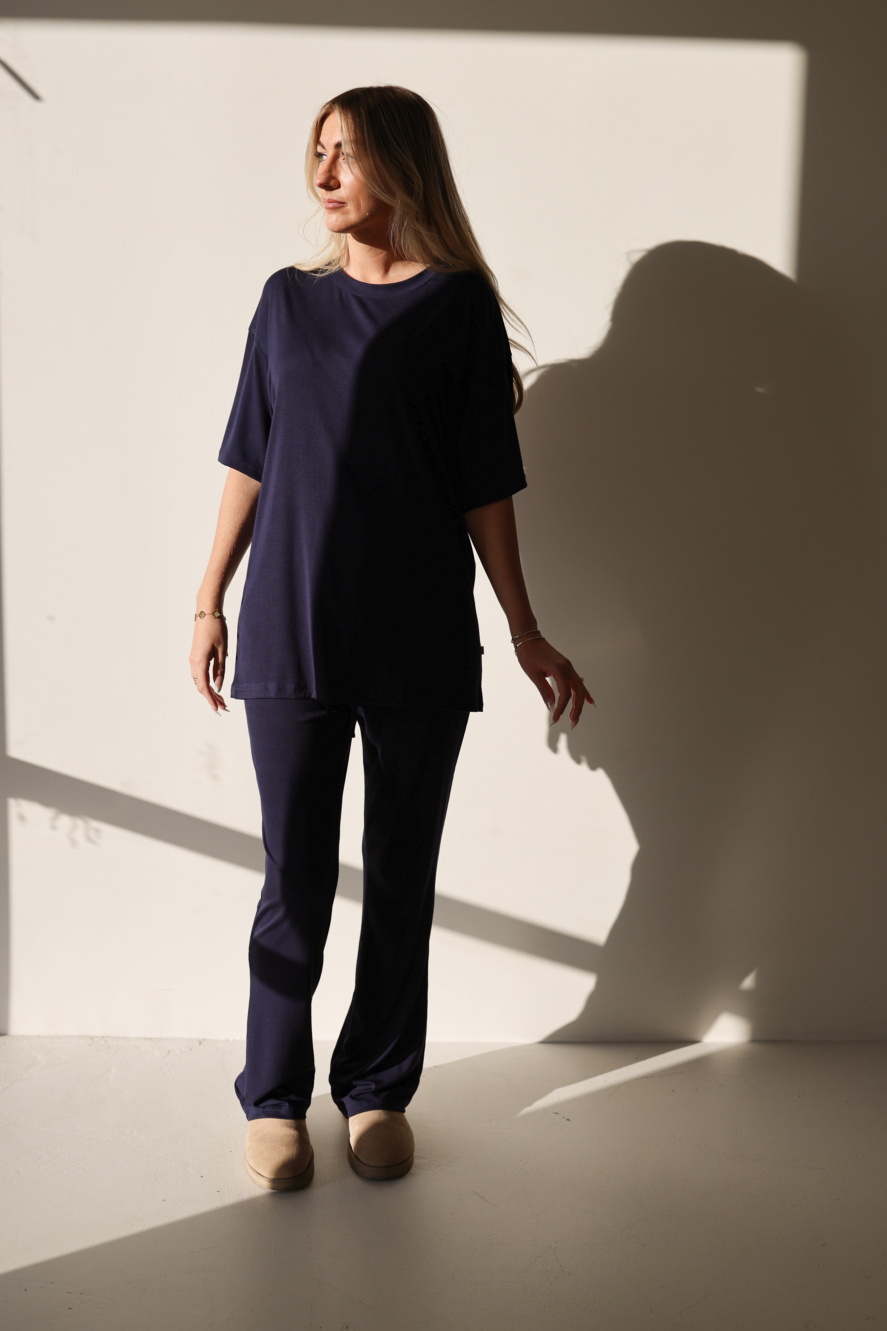 A woman wearing an oversized Blueberry Ribbed bamboo short sleeve top and matching flare pants while standing against a neutral wall.