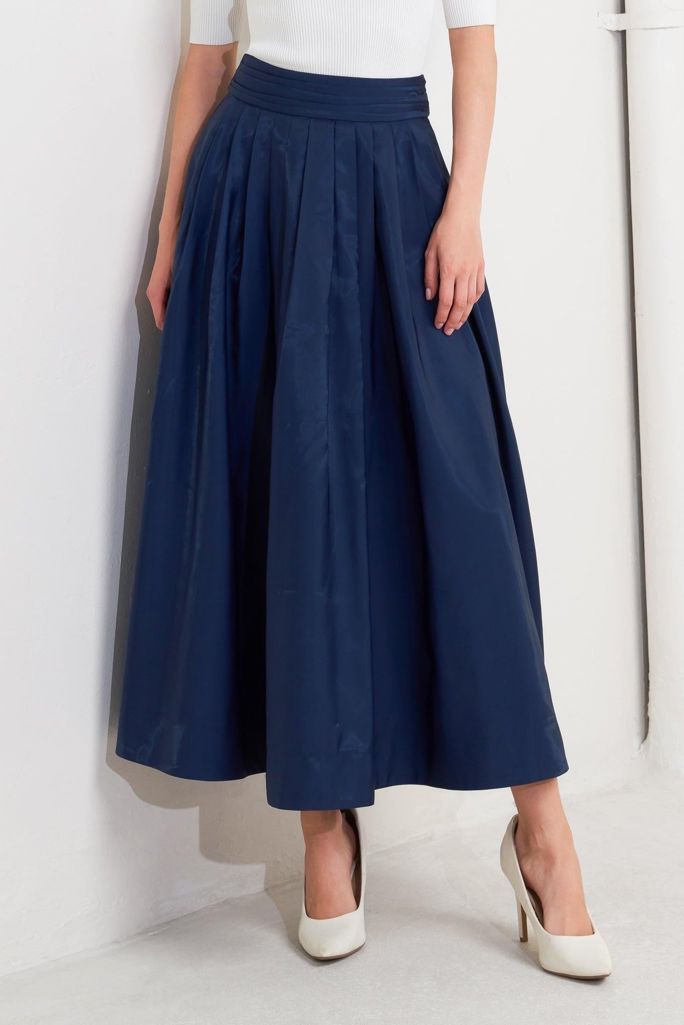 A woman wearing a navy woven pleated midi skirt with a visible back zipper closure, paired with white heels.